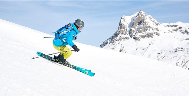 Ein Skifahrer fährt auf einer schneebedeckten Piste. Im Hintergrund sind beeindruckende Berge zu sehen.