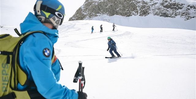 A ski instructor observes students on a snowy slope. In the background, there are other skiers and majestic mountains visible.