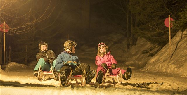 Drei Kinder sitzen auf Schlitten im Schnee bei Nacht. Es gibt Lichter und einige Wegmarkierungen im Hintergrund.