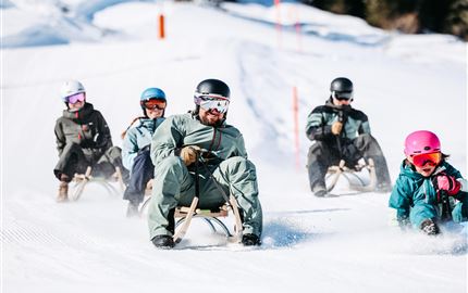 Eine Gruppe von Menschen fährt mit Schlitten eine verschneite Piste hinunter. Alle tragen Winterbekleidung und Helme, während sie Spaß im Schnee haben.