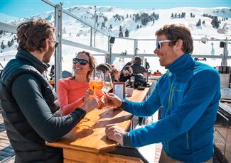 Drei Personen genießen Getränke auf einer sonnigen Terrasse in den Bergen. Im Hintergrund sind schneebedeckte Hänge zu sehen.