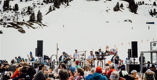 Eine lebendige Veranstaltung in den Bergen mit einer Band, die live spielt. Im Hintergrund sieht man Schnee und Skifahrer, während die Menschen in der Menge entspannen und feiern.