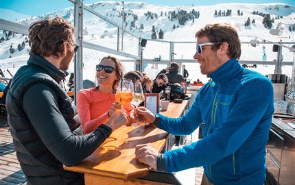 Three people are enjoying drinks on a sunny terrace in the mountains. In the background, snow-covered slopes can be seen.