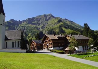 Eine malerische Alpenlandschaft mit grünen Bergen im Hintergrund. Im Vordergrund sind traditionelle Holzhäuser und eine Kirche zu sehen.