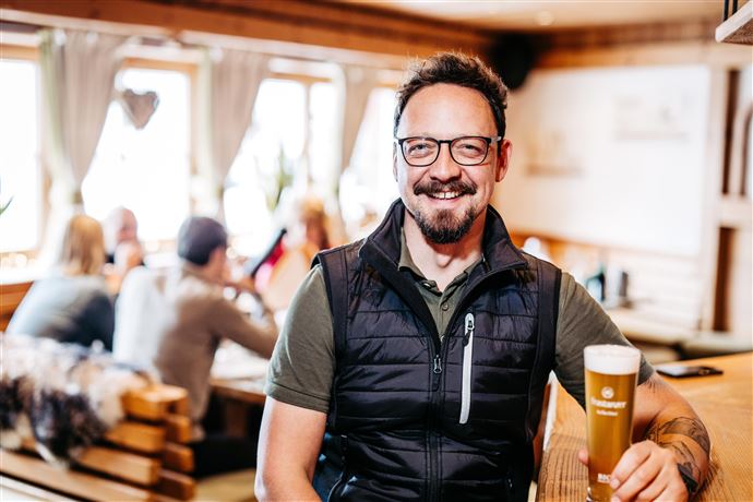 A man with glasses and a beard is sitting in a cozy restaurant, holding a glass of beer. In the background, other guests and an inviting atmosphere can be seen.