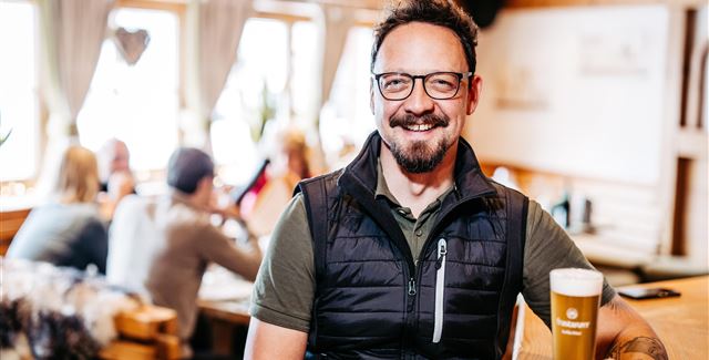 Ein Mann mit Brille und Bart sitzt in einem gemütlichen Restaurant und hält ein Glas Bier. Im Hintergrund sind andere Gäste und eine einladende Atmosphäre zu sehen.