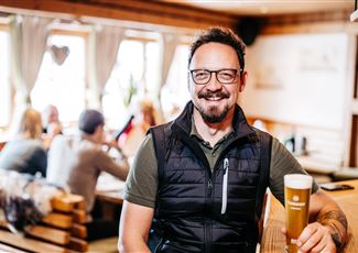A man with glasses and a beard is sitting in a cozy restaurant, holding a glass of beer. In the background, other guests and an inviting atmosphere can be seen.