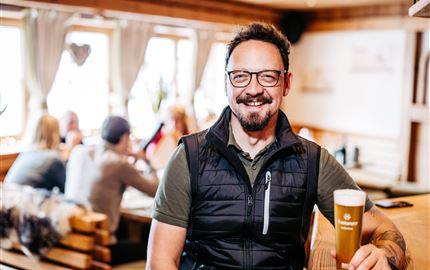 Ein Mann mit Brille und Bart sitzt in einem gemütlichen Restaurant und hält ein Glas Bier. Im Hintergrund sind andere Gäste und eine einladende Atmosphäre zu sehen.