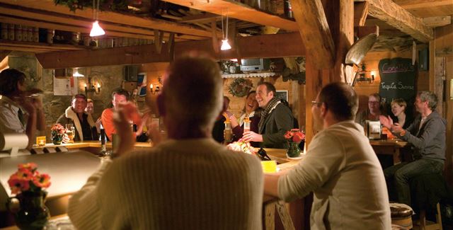 Eine gemütliche Bar mit Holzmöbeln und warmem Licht. Menschen sitzen an der Theke und haben eine gute Zeit miteinander.