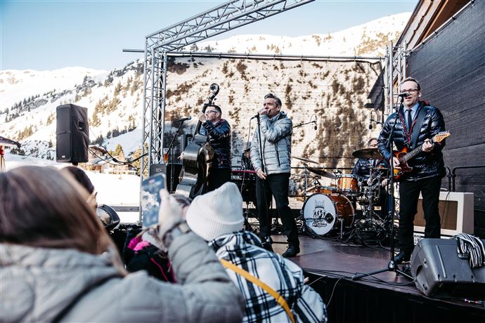 Eine Live-Band spielt auf einer Bühne in den Bergen. Im Hintergrund sind schneebedeckte Gipfel und Zuschauer, die das Konzert genießen.