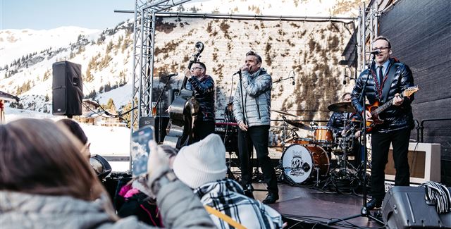 Eine Live-Band spielt auf einer Bühne in den Bergen. Im Hintergrund sind schneebedeckte Gipfel und Zuschauer, die das Konzert genießen.