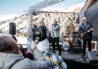 Eine Live-Band spielt auf einer Bühne in den Bergen. Im Hintergrund sind schneebedeckte Gipfel und Zuschauer, die das Konzert genießen.