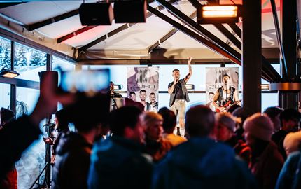 Eine lebhafte Veranstaltung in einer modernen Halle mit einer Band auf der Bühne. Die Menge jubelt und genießt die Musik.