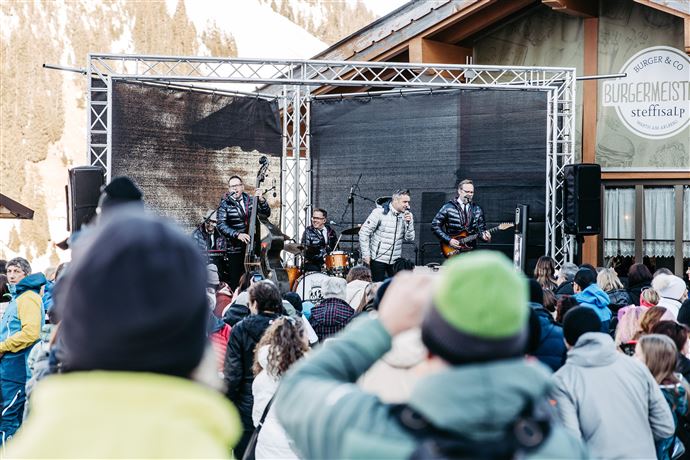 Eine Live-Band spielt auf einer Bühne in den Bergen. Im Hintergrund sind schneebedeckte Gipfel und Zuschauer, die das Konzert genießen.