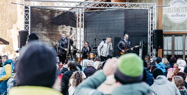 Eine Live-Band spielt auf einer Bühne in den Bergen. Im Hintergrund sind schneebedeckte Gipfel und Zuschauer, die das Konzert genießen.