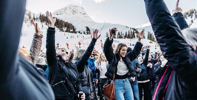 Eine Live-Band spielt auf einer Bühne in den Bergen. Im Hintergrund sind schneebedeckte Gipfel und Zuschauer, die das Konzert genießen.