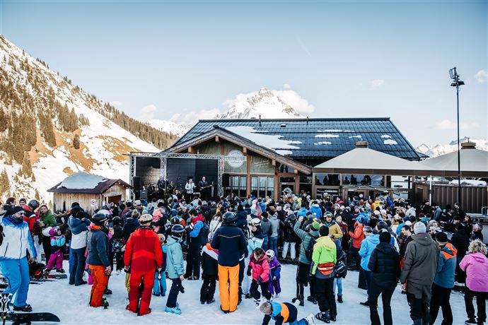 Eine Live-Band spielt auf einer Bühne in den Bergen. Im Hintergrund sind schneebedeckte Gipfel und Zuschauer, die das Konzert genießen.
