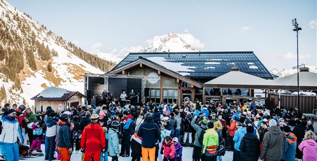 Eine Live-Band spielt auf einer Bühne in den Bergen. Im Hintergrund sind schneebedeckte Gipfel und Zuschauer, die das Konzert genießen.