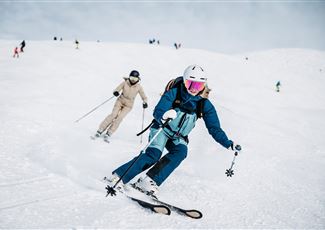 Eine Skifahrerin fährt den Hang hinunter, während eine weitere Person im Hintergrund fährt. Der Schnee ist frisch und die Landschaft wirkt winterlich.