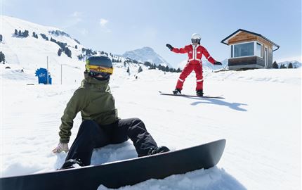 Eine Person fährt mit Skiern durch eine verschneite Landschaft. Im Hintergrund sind Berge und ein klarer blauer Himmel zu sehen.