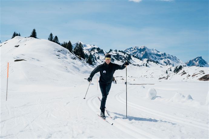 A person is skiing through a snowy landscape. In the background, mountains and a clear blue sky can be seen.