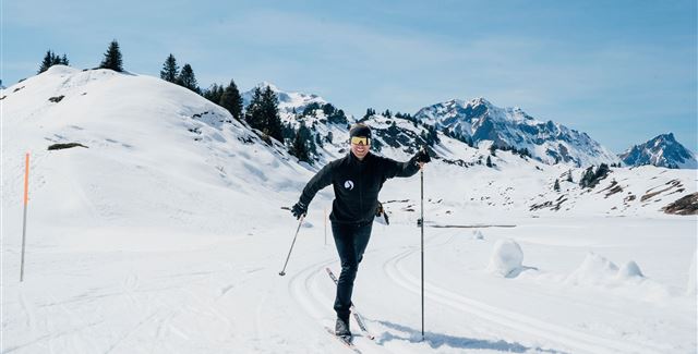 Eine Person fährt mit Skiern durch eine verschneite Landschaft. Im Hintergrund sind Berge und ein klarer blauer Himmel zu sehen.