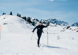 A person is skiing through a snowy landscape. In the background, mountains and a clear blue sky can be seen.