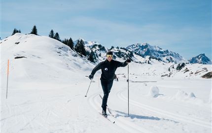 Eine Person fährt mit Skiern durch eine verschneite Landschaft. Im Hintergrund sind Berge und ein klarer blauer Himmel zu sehen.