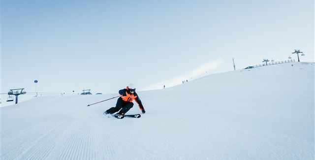 A skier is skiing on a snow-covered slope. The sky is clear and blue, and ski lifts are visible in the background.