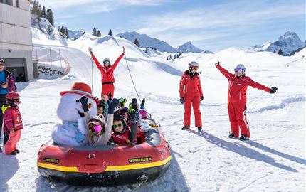 Eine Gruppe von Kindern in roten Skianzügen fährt in einem aufblasbaren Boot auf dem Schnee. Im Hintergrund sind schneebedeckte Berge und ein klarer blauer Himmel zu sehen.