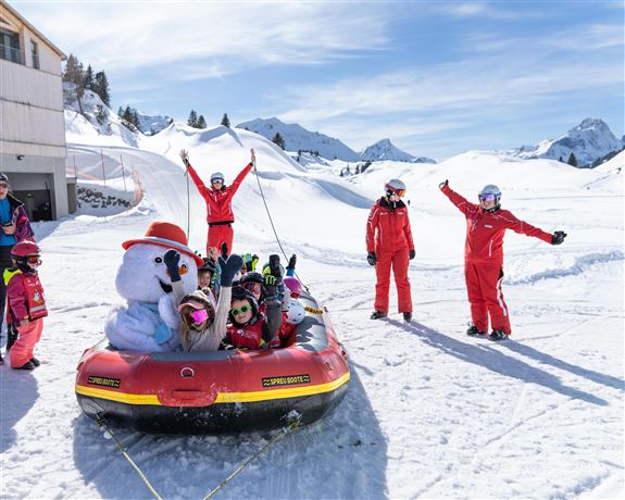 Eine Gruppe von Kindern in roten Skianzügen fährt in einem aufblasbaren Boot auf dem Schnee. Im Hintergrund sind schneebedeckte Berge und ein klarer blauer Himmel zu sehen.