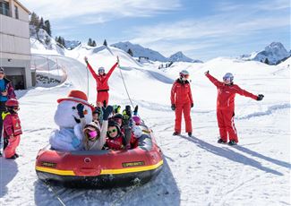 Eine Gruppe von Kindern in roten Skianzügen fährt in einem aufblasbaren Boot auf dem Schnee. Im Hintergrund sind schneebedeckte Berge und ein klarer blauer Himmel zu sehen.