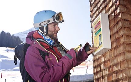 Ein Skilehrer beobachtet Schüler auf einer verschneiten Piste. Im Hintergrund sind weitere Skifahrer und majestätische Berge zu sehen.