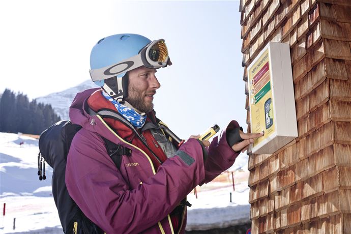 A ski instructor observes students on a snowy slope. In the background, there are other skiers and majestic mountains visible.