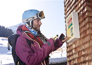 A ski instructor observes students on a snowy slope. In the background, there are other skiers and majestic mountains visible.