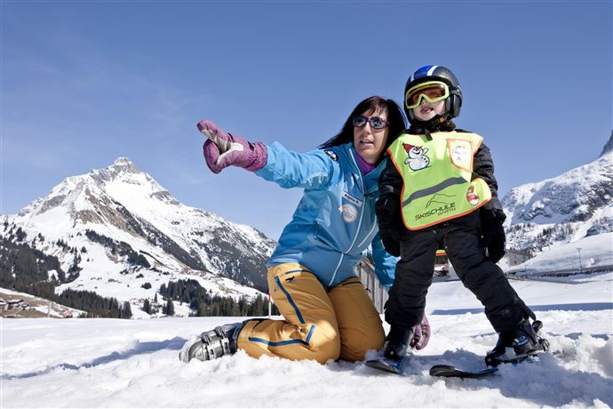 A ski instructor observes students on a snowy slope. In the background, there are other skiers and majestic mountains visible.