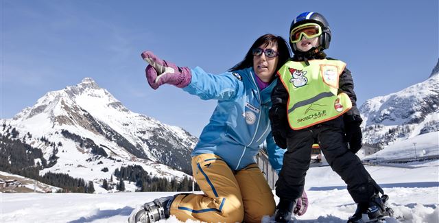 A ski instructor observes students on a snowy slope. In the background, there are other skiers and majestic mountains visible.
