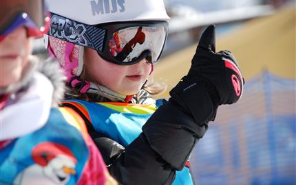 Ein Skilehrer beobachtet Schüler auf einer verschneiten Piste. Im Hintergrund sind weitere Skifahrer und majestätische Berge zu sehen.
