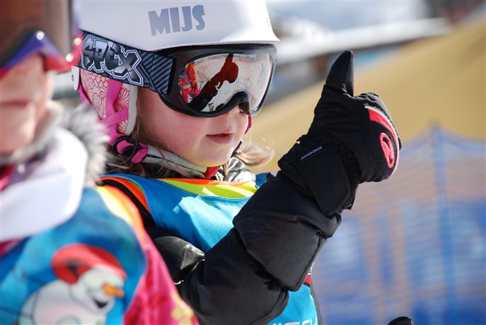 A ski instructor observes students on a snowy slope. In the background, there are other skiers and majestic mountains visible.