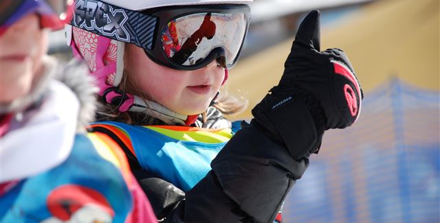 A ski instructor observes students on a snowy slope. In the background, there are other skiers and majestic mountains visible.