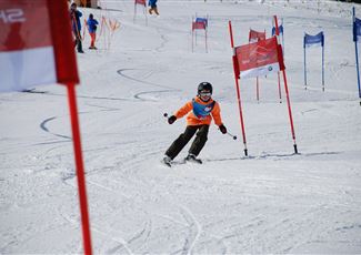 A ski instructor observes students on a snowy slope. In the background, there are other skiers and majestic mountains visible.