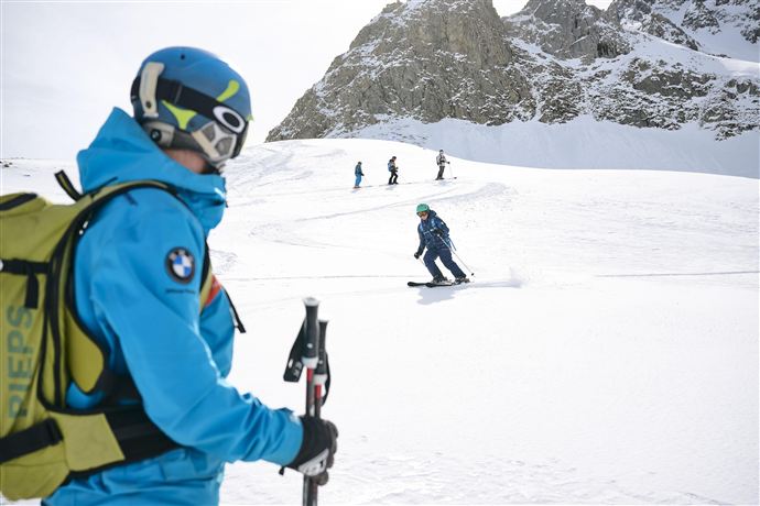 Ein Skilehrer steht in der verschneiten Berglandschaft und beobachtet Schüler beim Skifahren. Die Umgebung ist von hohen Bergen und klarem Himmel umgeben.