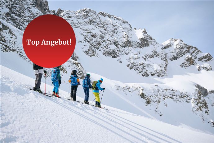 Eine Gruppe von Skifahrern steht auf einem verschneiten Hang, umgeben von majestätischen Bergen. Im Vordergrund steht ein roter Kreis mit der Aufschrift „Top Angebot!“.