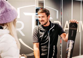 A ski expert is speaking with a customer and holding a pair of skis in hand. In the background, a stylish sports store can be seen.