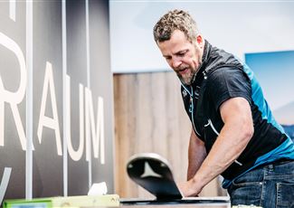Ein Mann arbeitet an einem Tisch und bereitet etwas vor. Im Hintergrund ist ein großes Schild mit dem Wort "RAUM" zu sehen.