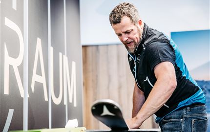Ein Mann arbeitet an einem Tisch und bereitet etwas vor. Im Hintergrund ist ein großes Schild mit dem Wort "RAUM" zu sehen.