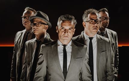 Eine Live-Band spielt auf einer Bühne in den Bergen. Im Hintergrund sind schneebedeckte Gipfel und Zuschauer, die das Konzert genießen.