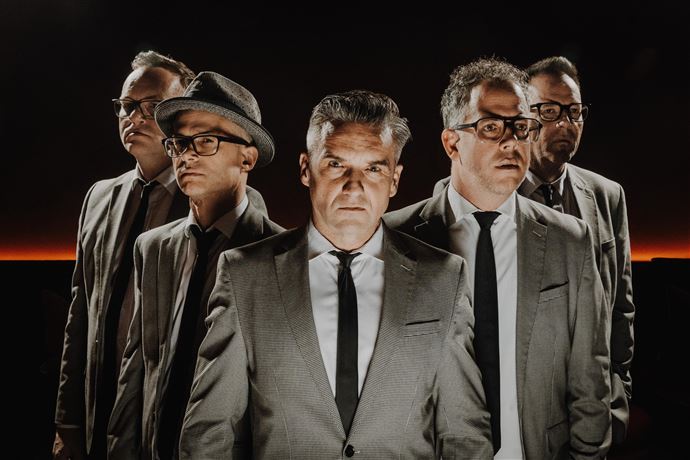 A live band is playing on a stage in the mountains. In the background, there are snow-covered peaks and spectators enjoying the concert.