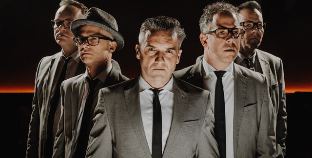 A live band is playing on a stage in the mountains. In the background, there are snow-covered peaks and spectators enjoying the concert.