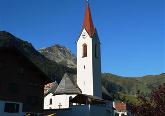 Eine Kirche mit einem spitzen roten Dach steht vor einer malerischen Berglandschaft. Im Vordergrund blühen gelbe Blumen.